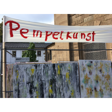 Banner mit der roten Aufschrift Pempelkunst über mehreren abstrakt bemalten Tafeln bei einer Ausstellung im Freien.