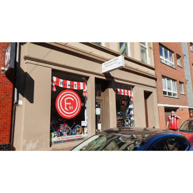 Schaufenster eines Gebäudes mit rot-weißer Markise und einem großen rot-weißen runden Emblem; im unteren Bereich sind gestaltete Objekte zu 