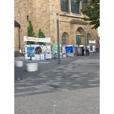 Auf einem gepflasterten Platz vor einem historischen Steingebäude sind mehrere farbenfrohe Bilder und Ausstellungstafeln aufgestellt.